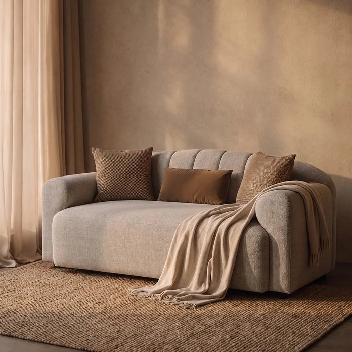 A modern beige upholstered loveseat sofa with three matching throw pillows and a draped cream throw blanket, set on a woven rug against a textured warm beige wall with curtains.