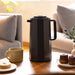 Black thermos on a wooden table with a cup, pastries, and a plant in a blurred living room setting.