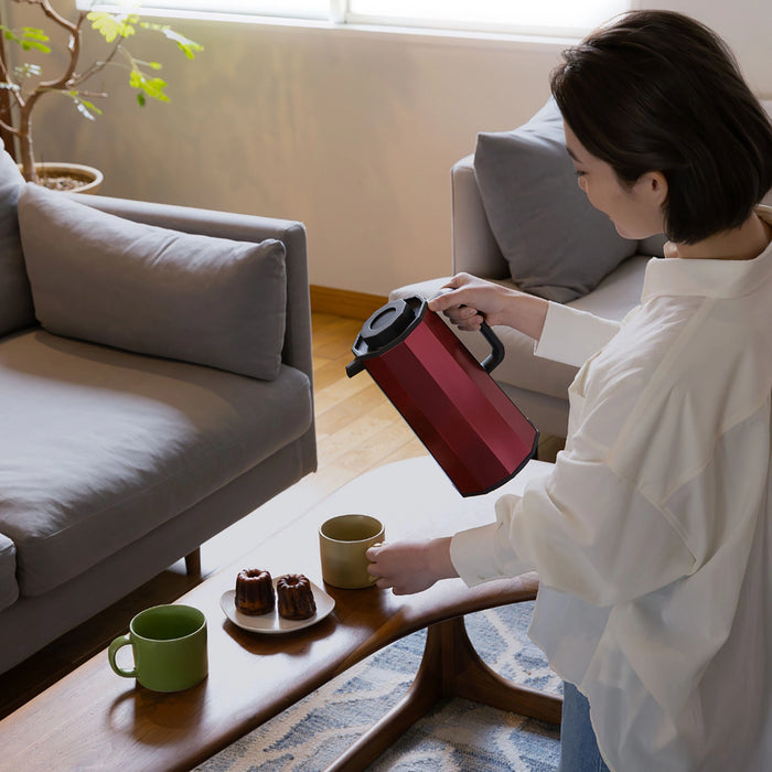 Woman pouring tea from a red thermos into a green mug on a coffee table in a living room.