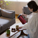 Woman pouring tea from a red thermos into a green mug on a coffee table in a living room.