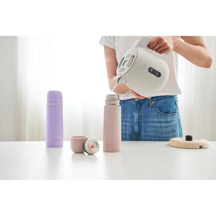Person pouring water from a white electric kettle into a pink thermos on a white surface.