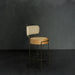 Bar stool with beige cushioned seat and backrest against a dark gray wall.