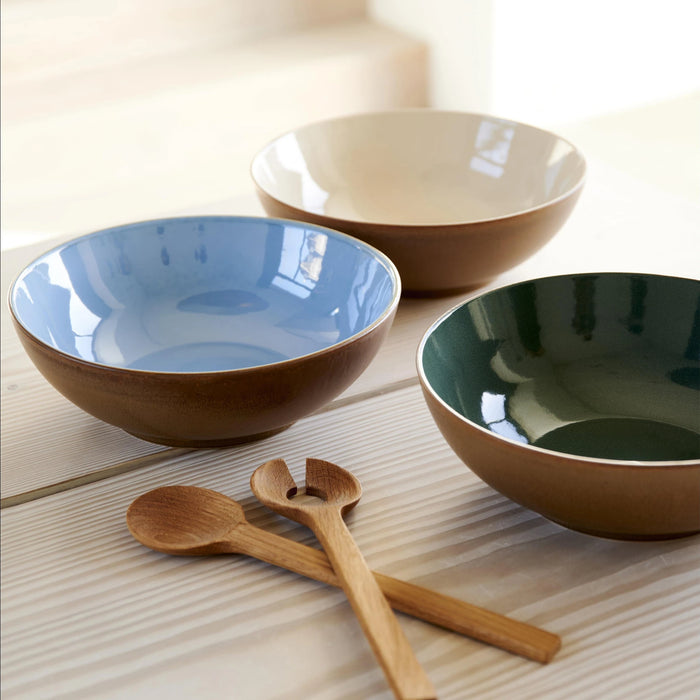 Three ceramic bowls in blue, beige, and green with wooden spoons on a light wooden surface.