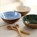 Three ceramic bowls in blue, beige, and green with wooden spoons on a light wooden surface.