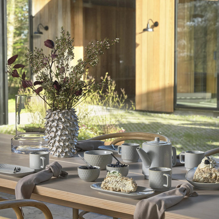 Outdoor dining table set with ceramic dishes, teapot, and flowers on a wooden deck.