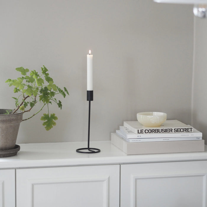 Candle in a black holder on a white surface with books and a plant in the background.