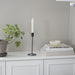 Candle in a black holder on a white surface with books and a plant in the background.
