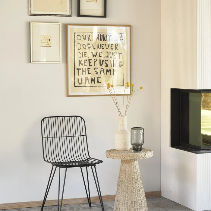 Modern interior with a black wire chair, woven side table, and framed artwork on a light wall.