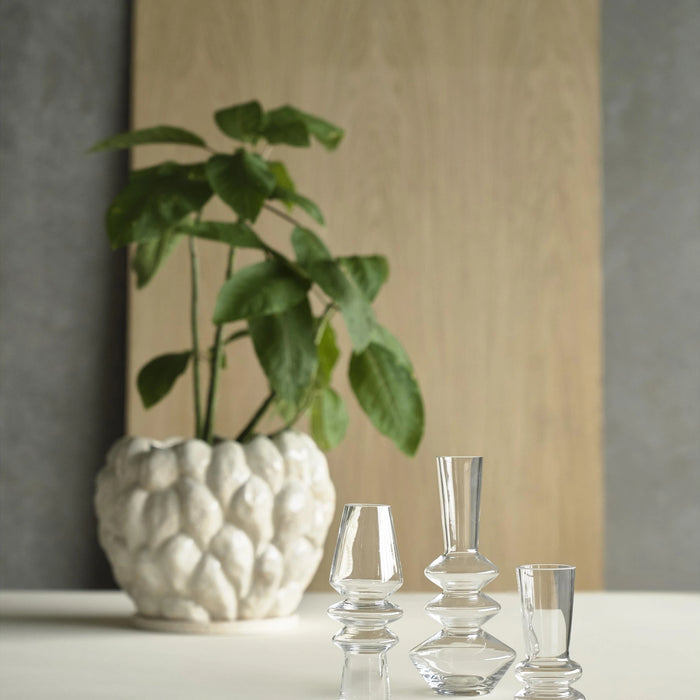 Set of clear glass vases on a surface with a textured white pot and green plant in the background.