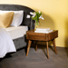 Wooden side table with a vase of flowers and books next to a bed with gray upholstered headboard.