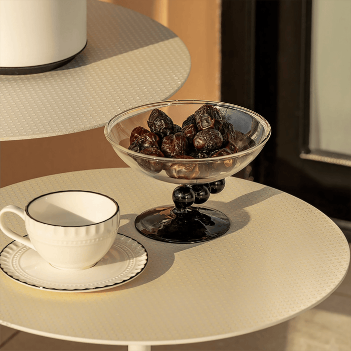 White teacup and saucer with a glass bowl containing dates on a beige table.