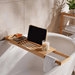Wooden bathtub caddy with tablet, glass of wine, and candle in a bathroom setting.