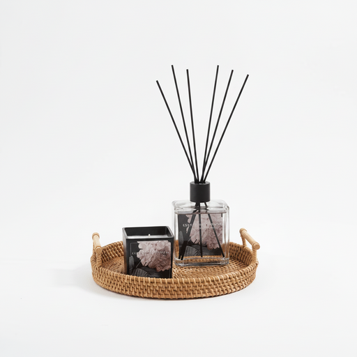 Diffuser with black sticks on a woven tray with a candle on a white background