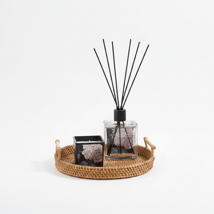 Diffuser with black sticks on a woven tray with a candle on a white background