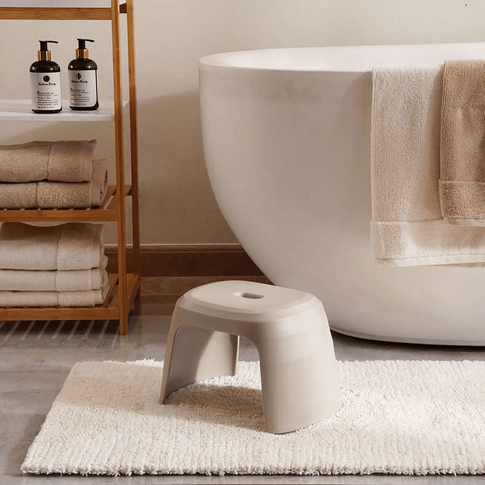 White stool in front of a bathtub with towels and bottles on a shelf in a bathroom setting.