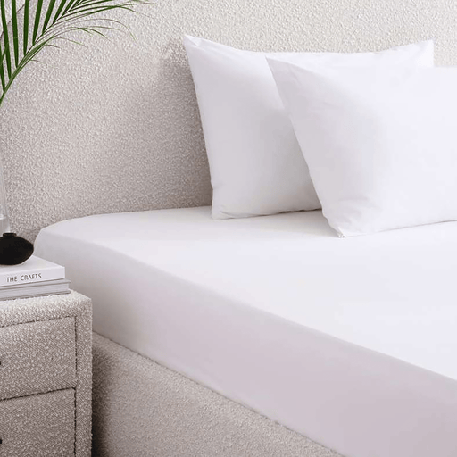 White bed sheets and pillows on a neatly made bed with a nightstand and plant in the background.
