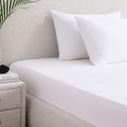 White bed sheets and pillows on a neatly made bed with a nightstand and plant in the background.
