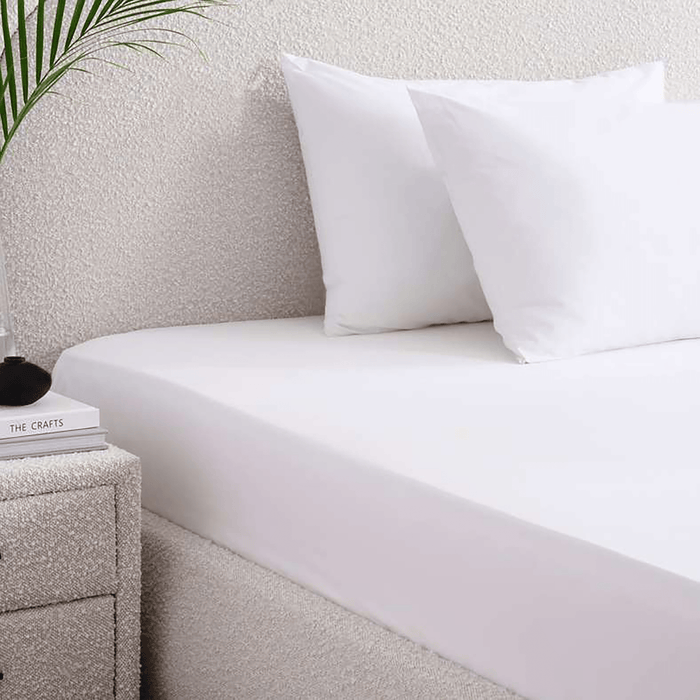 White bed sheets and pillows on a neatly made bed with a nightstand and plant in the background.