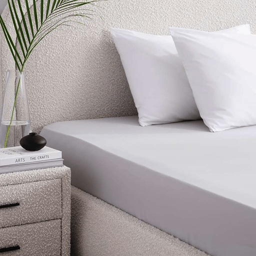 Bed with white bedding and pillows, nightstand with books and plant in the corner.