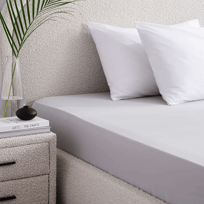 Bed with white bedding and pillows, nightstand with books and plant in the corner.