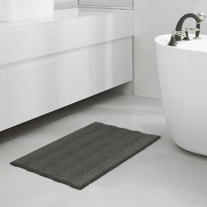 Gray bath mat on a bathroom floor with a white bathtub and sink in the background.