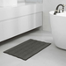 Gray bath mat on a bathroom floor with a white bathtub and sink in the background.