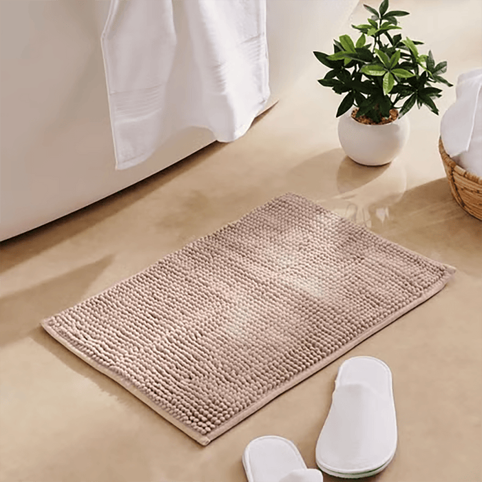 Beige bath mat on a bathroom floor with white slippers and a plant in the background.