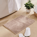 Beige bath mat on a bathroom floor with white slippers and a plant in the background.