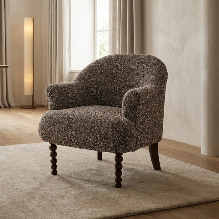 A single upholstered accent chair with a textured, speckled taupe-brown fabric and dark turned wooden legs, set on a cream rug in a softly lit living room.