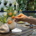 Hand holding a white outdoor lamp on a wooden table with plants and flowers in the background