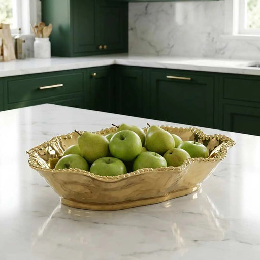A gold-toned scalloped metal bowl filled with green apples, placed on a white marble countertop in a modern kitchen with dark green cabinets.
