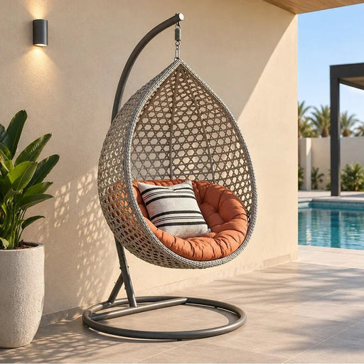 Teardrop-shaped woven outdoor hanging chair with an orange cushion and striped pillow, mounted on a curved metal stand on a patio beside a pool.