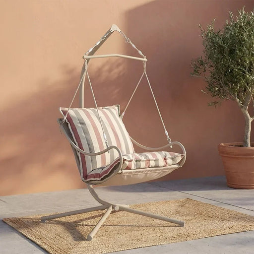 A hanging swing chair with a cream metal frame and striped cushions in beige, pink, and white, set on a woven rug against a warm peach wall with a potted olive tree nearby.