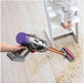 Person using a Dyson vacuum cleaner on a wooden floor with a child in the background.