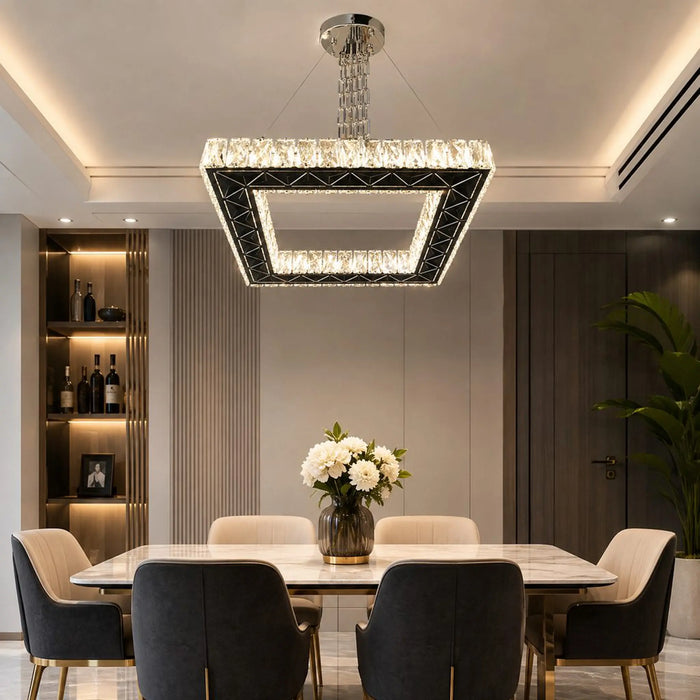 A modern square LED crystal chandelier hanging over a dining table in a contemporary dining room. The chandelier features a black geometric frame with embedded clear crystals and a glowing white LED light.