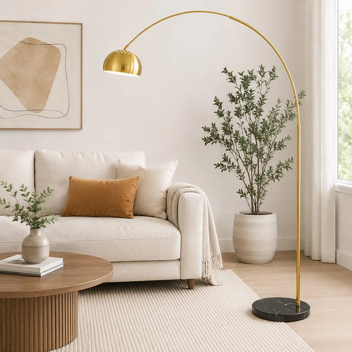 A modern gold arched floor lamp with a black marble base stands in a bright, contemporary living room next to a white sofa and a large potted plant.