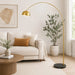 A modern gold arched floor lamp with a black marble base stands in a bright, contemporary living room next to a white sofa and a large potted plant.
