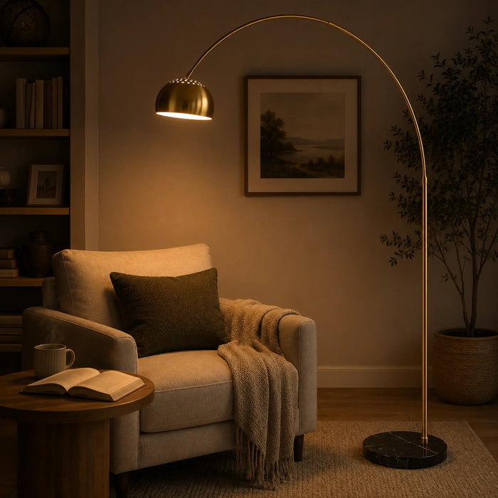 A tall arched floor lamp with a brushed gold finish stands in a warmly lit living room beside a beige armchair, with its light turned on.