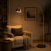 A tall arched floor lamp with a brushed gold finish stands in a warmly lit living room beside a beige armchair, with its light turned on.
