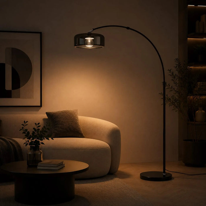 A modern black arched floor lamp with a brushed metal shade, glowing warmly in a dimly lit living room next to a beige sofa.