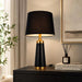 A modern table lamp with a black fabric drum shade, a black tapered base, and gold metal accents, shown lit on a dark wooden side table in a styled interior setting.