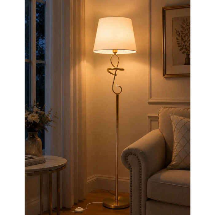 A classic brass floor lamp with a warm glowing white drum shade, styled in a cozy living room next to a beige armchair and a window.
