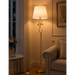 A classic brass floor lamp with a warm glowing white drum shade, styled in a cozy living room next to a beige armchair and a window.