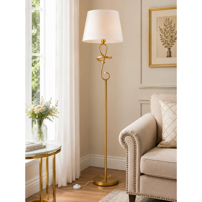 A tall floor lamp with a white drum shade and ornate gold metal frame stands in a bright living room beside a beige armchair and a small side table.