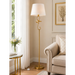 A tall floor lamp with a white drum shade and ornate gold metal frame stands in a bright living room beside a beige armchair and a small side table.
