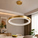A modern circular pendant light with a brushed gold metal frame and white LED inner ring, hanging from a ceiling in a stylishly decorated dining room.