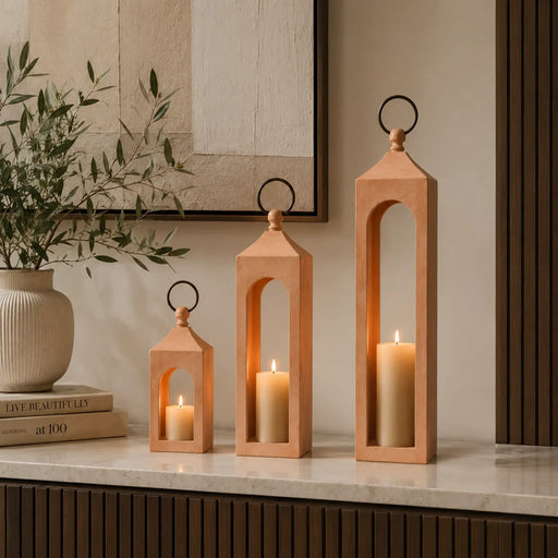 Set of three wooden lanterns with candles on a surface, with a plant and books in the background.