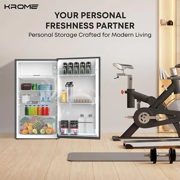 Krome refrigerator with open door displaying food and drinks, next to a stationary bike in a room setting.