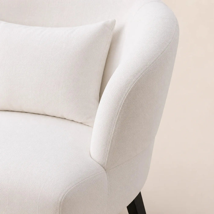 Close-up of a cream-colored upholstered armchair with a matching throw pillow, showing the textured fabric and curved armrest against a light beige background.