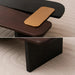 A modern low wooden table with a matte dark brown wood finish and a contrasting black top panel, shown with a gold rectangular object resting on it. The bottom half shows the table leg on a textured beige carpet.